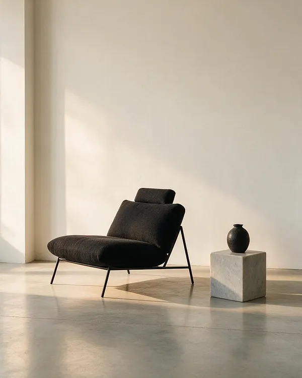 A minimalist room with a sculptural black upholstered lounge chair on thin metal legs, placed beside a small marble cube pedestal holding a matte black vase. The space has pale walls, a polished concrete floor, and soft sunlight creating gentle shadows.