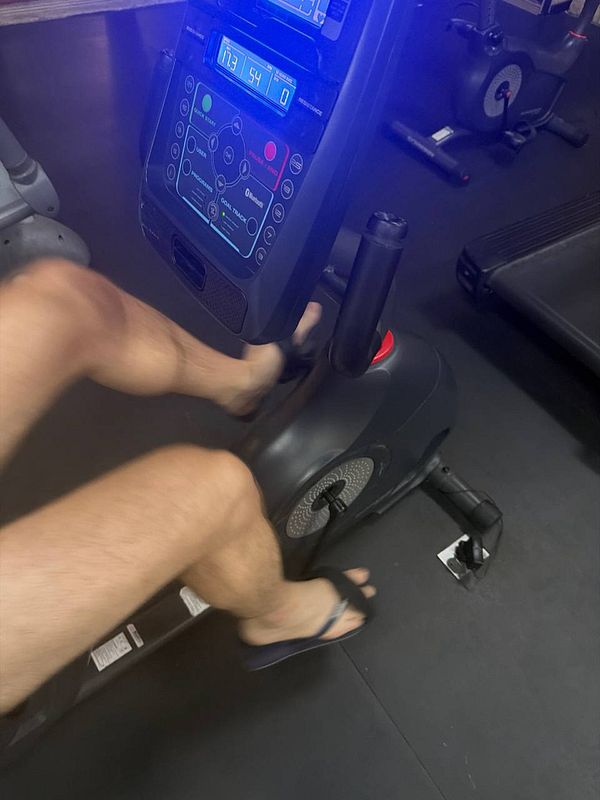 A woman wearing bright pink workout leggings and a black top is performing a weighted dumbbell row exercise in a gym with a stone wall and shiny floor.