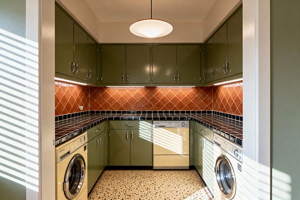 A U-shaped mid-century modern laundry room with glossy avocado-green upper and lower cabinets and chrome pulls, black ceramic tile countertops, and a diamond-pattern terracotta backsplash lit by continuous under-cabinet lighting. A saucer pendant centers the space above cream appliances integrated under the counters, including two front-loading machines, while speckled terrazzo flooring and light walls reflect daylight filtering through blinds at the doorway.
