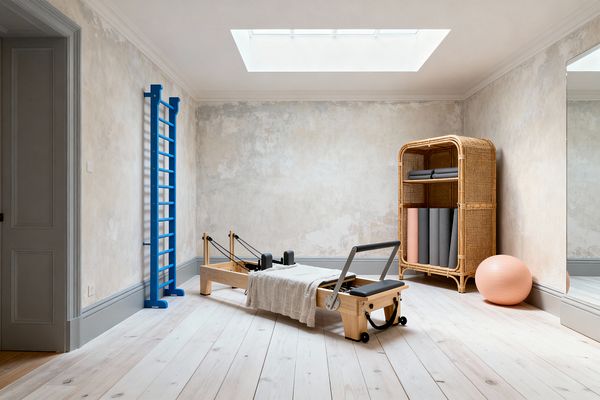 A serene home gym with shabby chic textures features a wooden Pilates reformer centered on pale whitewashed floorboards beneath a skylight. Distressed plaster walls with gray wainscotting frame a bright cobalt wall ladder on the left. To the right, a rattan cabinet stores rolled gray mats beside a peach exercise ball, with a tall mirror reflecting the room. Soft linen drapes over the reformer, and muted gray doors and trim complete the pared-back palette.