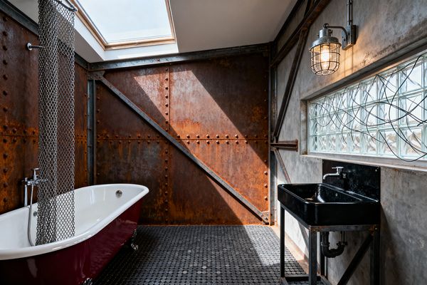 An industrial bathroom with riveted, rusted steel wall panels and exposed bracing, lit by a skylight. A freestanding clawfoot tub with a burgundy exterior and white interior sits on black penny-round floor tiles beside a hanging stainless chain shower curtain. On the right, a black enameled utility sink with exposed plumbing is mounted to a troweled concrete wall beneath a glass-block ribbon window and a galvanized caged sconce.