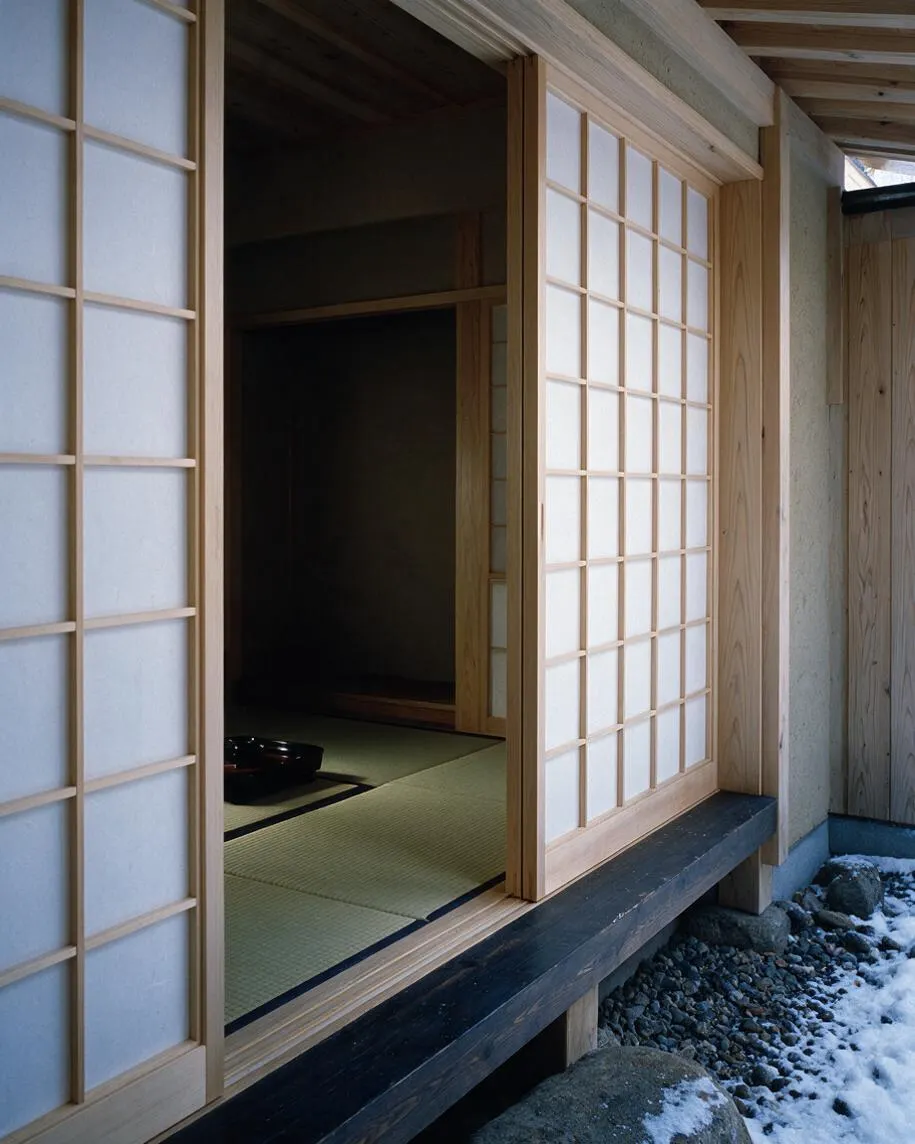 A Japanese Zen exterior showing a wooden facade with sliding shoji screens and an open entry revealing a dim tatami room inside. The structure sits above a dark base beam, with gravel, rocks, and patches of snow below, creating a calm, natural setting.
