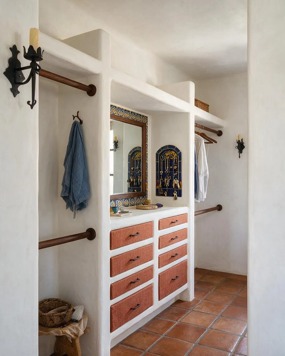 A Mediterranean-style closet with white stucco built-ins, terracotta tile flooring, and warm wood clothing rods. The center features a mirror framed with blue and yellow patterned tile and a small arched niche display, while woven drawer fronts add texture and earthy color. Black wall sconces and rustic accents complete the airy, handcrafted look.