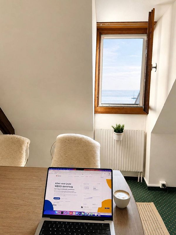A laptop on a wooden table in a rustic room with large windows showing green trees outside, providing a scenic view while working.