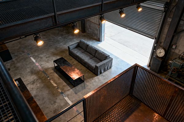 A double-height industrial living room viewed from an upper steel mezzanine shows a gray three-seat sofa paired with a long, low metal coffee table on a sealed concrete floor marked by a faded painted stripe. Warm spotlights hang from the catwalk while daylight pours through a partially open corrugated roll-up door. Exposed concrete block walls, a perforated steel railing with rust patina, visible conduit, and a large analog clock emphasize the raw warehouse character.
