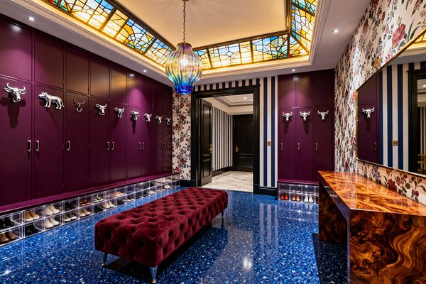 A maximalist mudroom with tall plum lockers fitted with chrome animal-head hooks above illuminated clear cubbies holding shoes. A deep burgundy tufted velvet bench sits on a glossy cobalt terrazzo floor. One wall mixes navy-and-white vertical stripes with large floral wallpaper, opposite a high-sheen burl wood console and brass-framed mirror. Overhead, stained-glass ceiling panels and an iridescent glass pendant cast warm, jewel-toned light; black doors with brass hardware lead out.