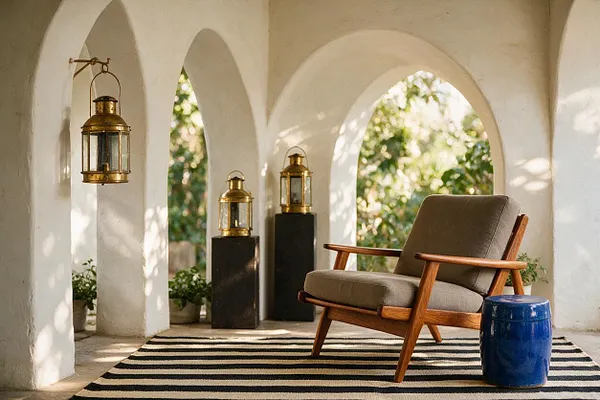 A sunlit outdoor porch with white stucco arches, brass lanterns on black pedestals, a striped rug, and a mid-century wooden lounge chair with gray cushions. A glossy blue ceramic stool adds a bold color accent, while greenery is visible through the arches in the background.