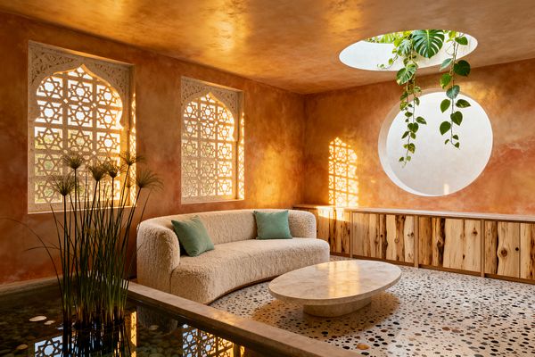 Sunlit living room with terracotta-toned plaster walls features two arched stone lattice windows and a circular skylight from which trailing vines hang. An ivory curved bouclé sofa with green cushions sits on a pebble-speckled terrazzo floor beside a shallow indoor reflecting pool planted with papyrus; a low oval travertine table and a long natural-wood credenza line the wall as patterned shadows warm the space.