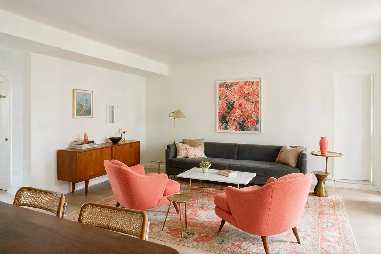 A bright eclectic living room with a charcoal sofa, two coral armchairs, and a pink patterned area rug. A mid-century wooden sideboard, brass side tables, and colorful floral wall art add warmth against the white walls.