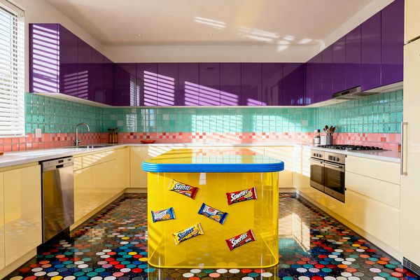 A vibrant Pop Art kitchen with glossy purple upper cabinets and buttery yellow lower cabinets around white quartz counters in a U-shape. A translucent yellow island with a rounded turquoise-edged top stands center, decorated with retro snack-wrapper magnets. Mint green square tiles with a pink checker stripe form the backsplash. The floor is a shiny black base covered in multicolored polka dots, with stainless appliances and daylight from a window with blinds.
