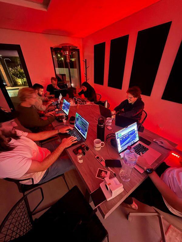 A group of six people working on laptops around a long table in a room with red lighting, collaborating in a coworking space.