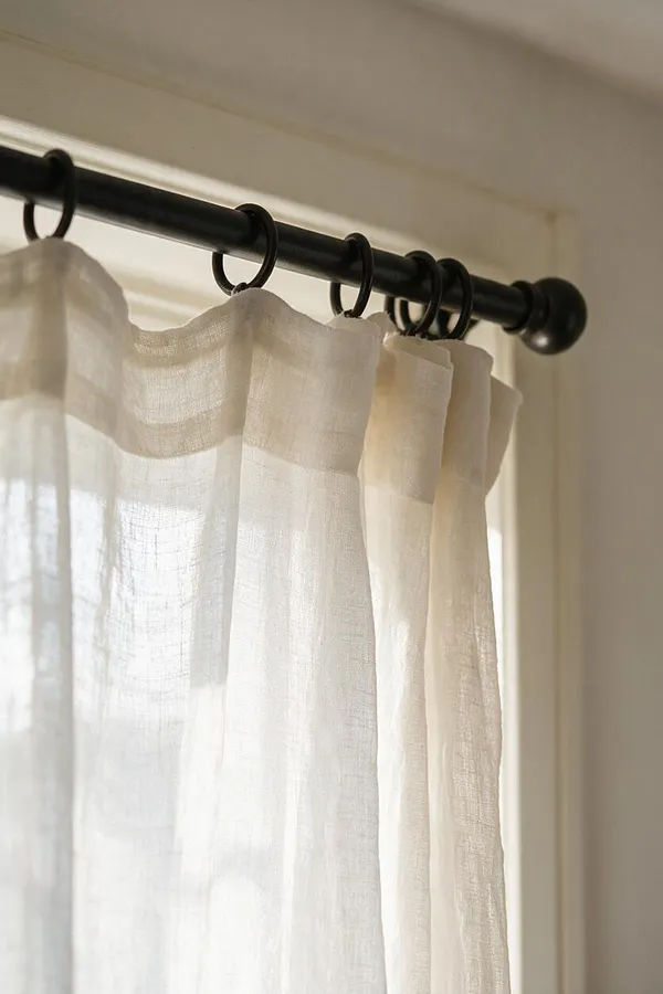 Close-up of soft ivory sheer curtains hanging from a matte black metal rod with circular rings beside a bright window. The lightweight fabric filters daylight, creating a calm Scandinavian look with minimal, airy finishes.
