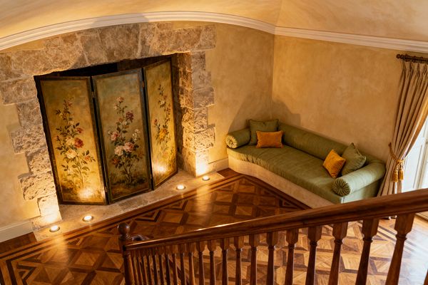 A cozy French Country media room seen from above shows a carved wood stair rail, geometric walnut-and-oak parquet flooring, and a large stone niche fitted with a hand-painted floral folding screen illuminated by small floor uplights. To the right, a long olive-green linen banquette with bolster arms and ochre pillows rests against warm beige plaster walls, beneath crown molding and beside tied-back drapery on a brass rod, creating an intimate, softly lit seating area.