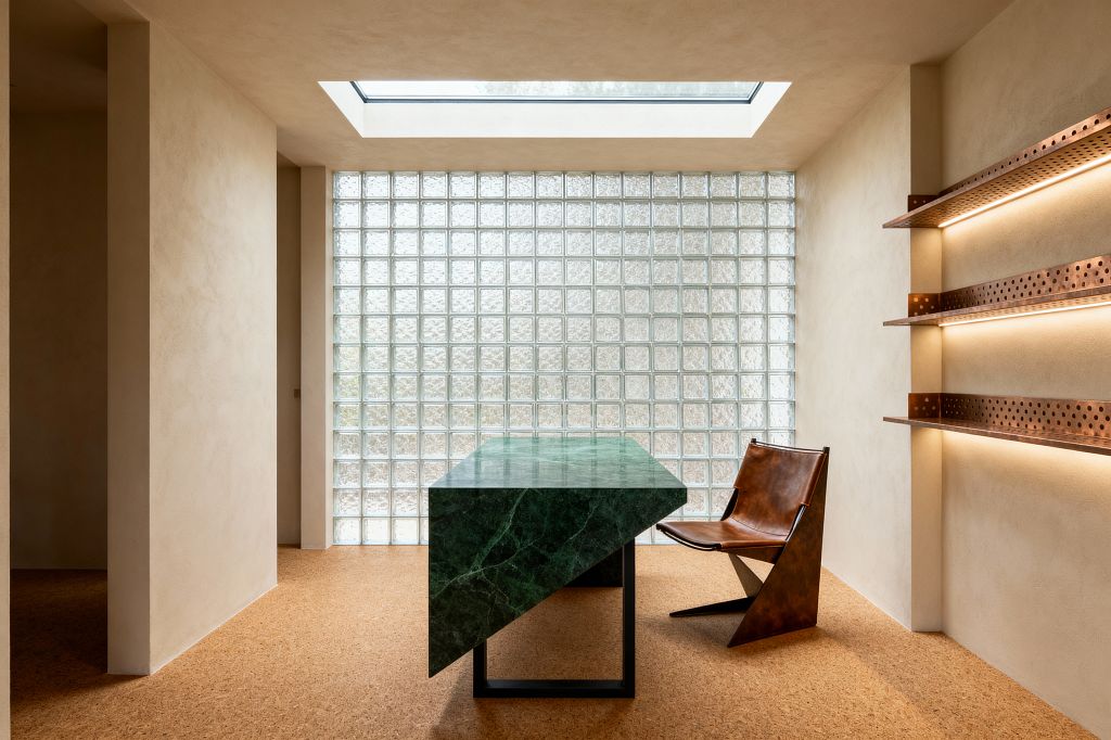 A contemporary home office centers on a sculptural emerald-green marble desk with an angular waterfall edge and a black steel support, positioned before a full-height glass-block wall. A roof skylight floods the room with daylight, highlighting warm lime-plaster walls and cork flooring. On the right, perforated copper shelves with integrated LED strips glow softly above a low cognac leather sling chair.