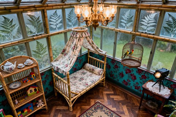 A children’s room styled like a Victorian conservatory features a rattan daybed with floral chintz mattress, bolster, and draped canopy beneath an ornate brass chandelier. Teal wallpaper with burgundy roses lines the lower walls, etched fern glass panels frame garden views, and a wicker shelf holds toys and a tea set; a hanging brass birdcage, lace-topped side table, vintage spotlight, and herringbone wood floor complete the scene.