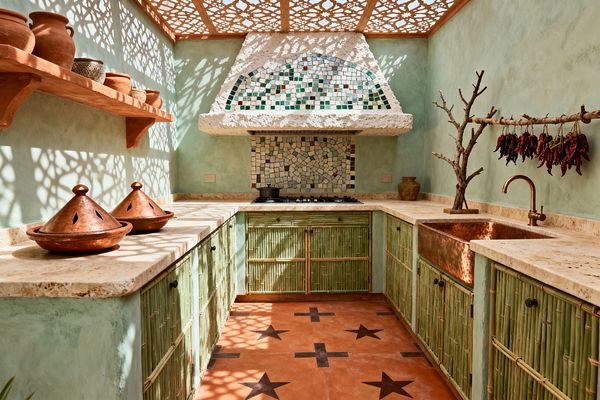 A compact U-shaped kitchen features sage green bamboo-front cabinets with cream travertine countertops and a hammered copper apron-front sink. A zellige mosaic backsplash climbs onto a textured hood beneath a carved wood lattice ceiling that casts patterned shadows; terracotta floor tiles with black star motifs anchor the space, while an open terracotta shelf displays pottery and tagines and a branch rack holds drying red chilies beside a centered gas cooktop.