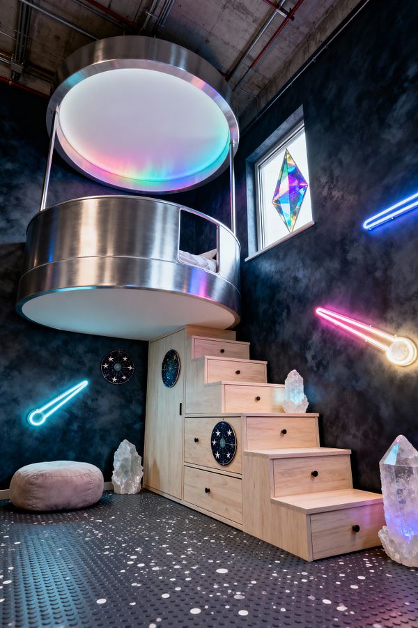 A kids’ room with a circular loft bed wrapped in brushed metal and a large diffused canopy that casts rainbow light. Pale wood stairs with storage drawers climb to the bed against charcoal, galaxy-effect walls with pink and blue neon comet lights. A dichroic prism hangs in the window, clear crystal sculptures sit on the steps and floor, a gray pouf rests nearby, and speckled black rubber coin flooring anchors the space beneath exposed ceiling pipes.