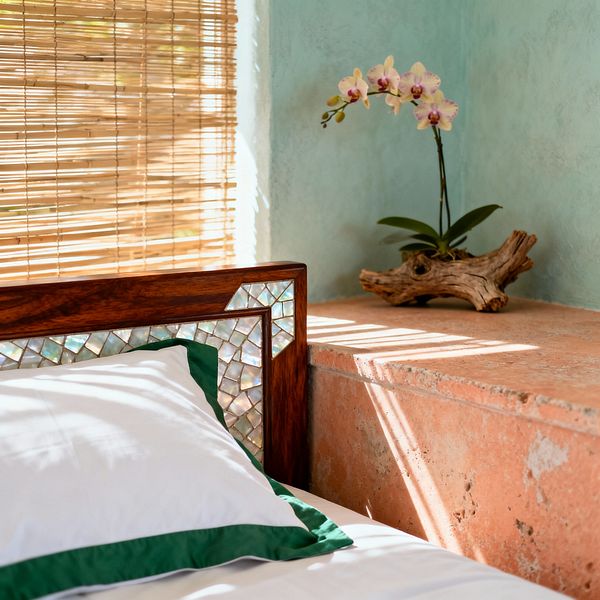 Close-up of a tropical bedroom corner with a mahogany wood headboard inlaid with shimmering mother-of-pearl tiles beside a coral-toned stone ledge. Sunlight filters through a bamboo roller blind onto a seafoam plaster wall, illuminating crisp white bedding with green trim and a small orchid displayed on a piece of driftwood.
