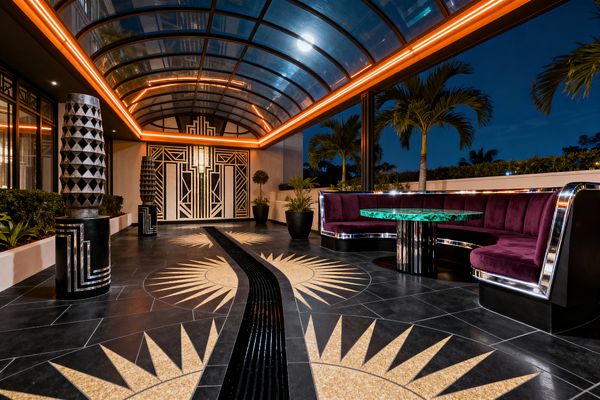Nighttime patio beneath a curved glass canopy edged with amber LED strips, leading to a geometric black-and-gold Art Deco doorway. The floor is black tile with gold terrazzo sunburst inlays and a wavy central channel. At right, a U-shaped plum velvet banquette with chrome trim surrounds a round green marble table on a black fluted base. Sculptural black-and-silver totems and black planters with tropical plants line the space, with palm trees visible beyond.