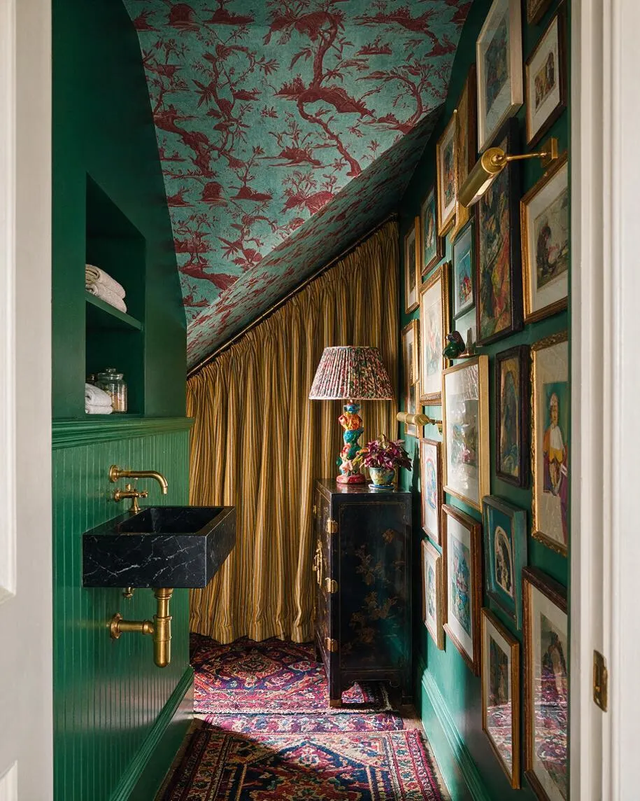 A narrow powder room with emerald green walls, a black marble wall-mounted sink, and brass plumbing and sconce. The ceiling is covered in turquoise and burgundy botanical wallpaper, one wall is filled with framed art, and gold drapery and patterned rugs add a richly layered maximalist look.