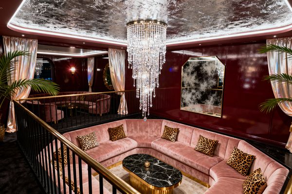 A sunken living room in vintage glam style features a curved blush-pink velvet sectional with leopard-print pillows surrounding a black marble coffee table on a fluted gold base. Glossy burgundy walls, champagne satin drapes, and potted palms frame the space. Overhead, a silver-leaf ceiling with cove lighting and a cascading crystal chandelier add a dramatic, sparkling glow.