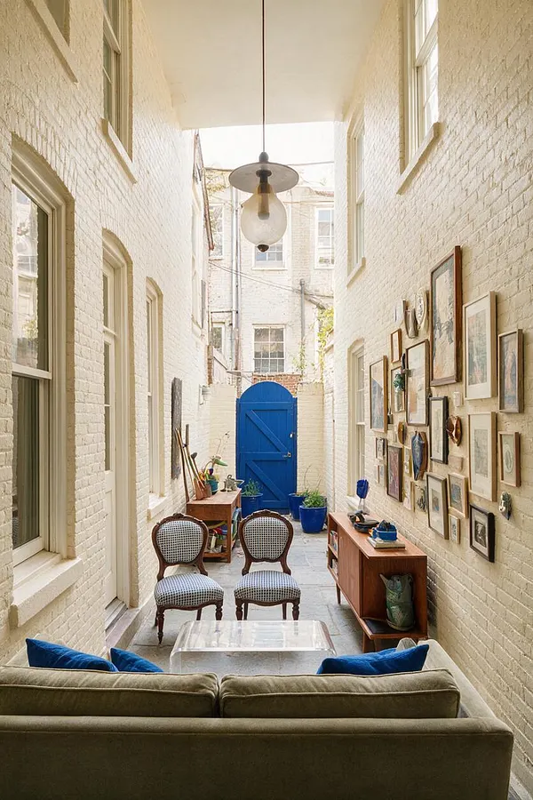 A narrow backyard courtyard with cream-painted brick walls, tall windows, and a bright blue gate at the far end. The space mixes eclectic furnishings including cane chairs, a sofa with blue pillows, a clear coffee table, wooden cabinets, hanging art, and potted plants beneath a simple pendant light.