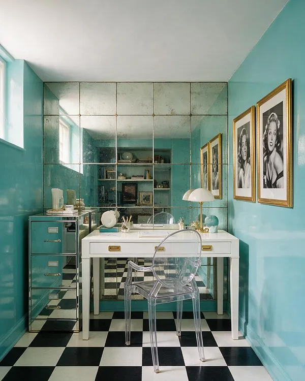 A compact basement vanity area painted in glossy turquoise, with a white desk centered beneath a large mirrored wall. Black-and-white checkered flooring, gold-framed black-and-white portraits, a clear acrylic chair, and a mirrored chest create a glamorous Hollywood Regency look.