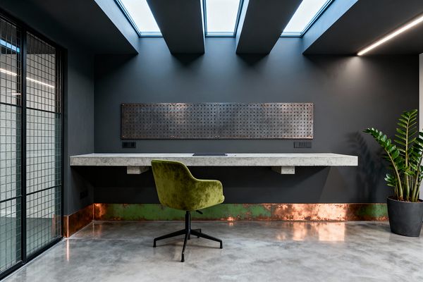 A minimalist industrial home office features a floating concrete desk on a dark charcoal wall beneath overhead skylights. A long perforated steel panel hangs above the work surface, while a band of patinated copper with green verdigris runs along the base of the wall. An olive green velvet swivel chair sits on polished concrete flooring. To the left, a black steel-framed glass partition with a grid pattern screens an adjacent space. A slim linear LED light and a potted plant add brightness.