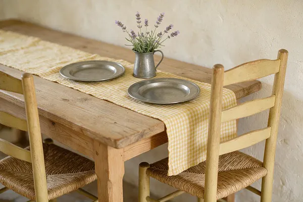 A simple French Country dining vignette with a rustic wooden table, two pale yellow chairs with woven seats, and a yellow gingham table runner. Two metal plates and a small pitcher of lavender sit on the table against a textured cream wall.