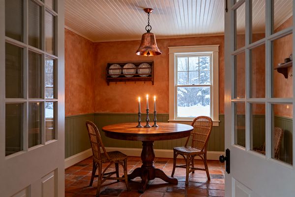 A cozy farmhouse dining room seen through white French doors features a round dark wood pedestal table with three lit metal candlesticks, flanked by two rattan chairs on terracotta tile flooring. Warm terracotta plaster walls sit above sage green beadboard wainscoting, a white beadboard ceiling, and a bell-shaped copper pendant. A wall plate rack holds white dishes, and a single window shows a snowy landscape outside.