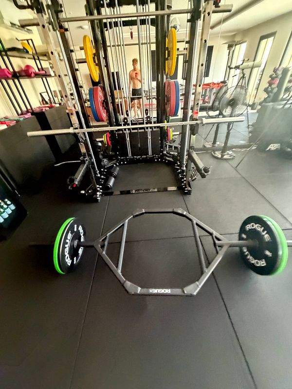 A muscular man lifting a trap bar loaded with plates inside a gym environment.