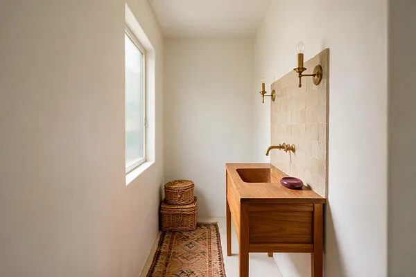 A narrow powder room with white walls, a frosted window on the left, and a warm wood vanity with an integrated sink on the right. A cream textured tile backsplash, brass wall sconces and faucet, woven baskets, and a patterned runner add bohemian character.