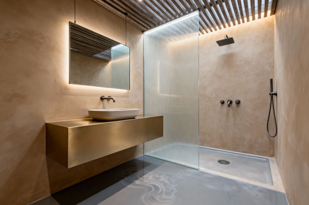 A modern bathroom with warm beige plaster walls features a floating brushed-brass vanity holding a pale stone vessel sink and a wall-mounted spout beneath a large, backlit mirror. A walk-in shower is defined by a reeded-glass screen, a ceiling-mounted square rain head, and matte black controls with a handshower; a slatted wood ceiling with concealed LEDs casts a soft glow over a seamless gray resin floor and integrated shower tray.