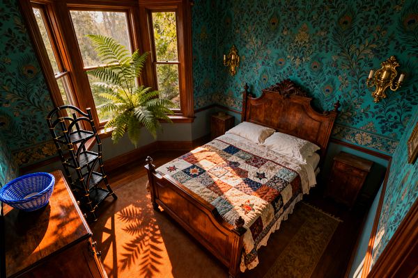 A Victorian bedroom with teal peacock-patterned wallpaper and gilded brass wall sconces frames a carved mahogany bed dressed with a multicolor patchwork quilt and white pillows. Sunlight streams through a bay window with deep wood trim, casting fern shadows across an orange-brown rug; a wooden nightstand, black étagère with gold accents, blue woven basket, and a narrow runner underline the room’s rich period details and warm atmosphere.