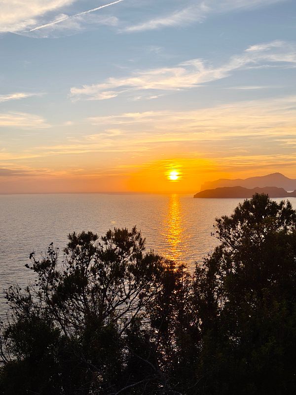 Sunset over calm water with silhouetted land and mountains in the distance under a dramatic sky.