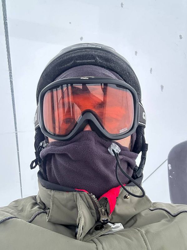Person holding a red ski with a snowboard on snowy terrain, wearing black gloves and blue jacket.