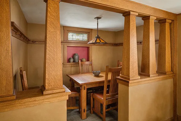 A Craftsman-style basement dining nook with thick wood columns, matching trim, and a square wooden table surrounded by slatted chairs. Warm tan walls, a red recessed display niche, and a stained-glass pendant light create an intimate, handcrafted feel.
