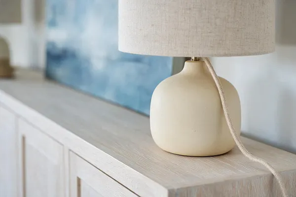 A neutral ceramic table lamp with a rounded cream base and textured linen shade sits on a light wood cabinet. A braided cord drapes down the side, and a blurred blue artwork in the background adds a calm coastal feel.