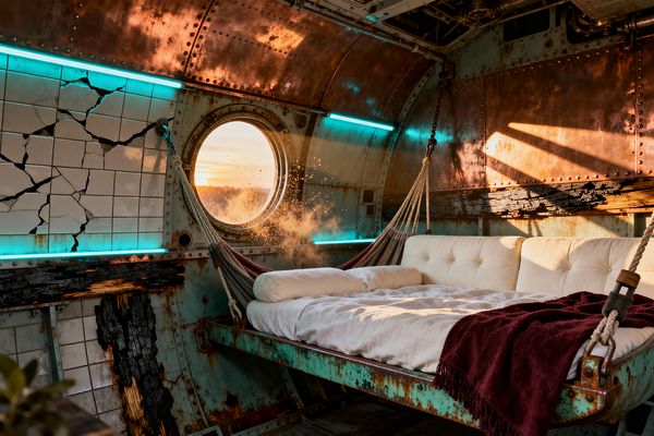 A bedroom set inside a corroded metal hull with riveted copper and teal-patinated steel panels, cracked white tiles highlighted by cyan tube lights, and exposed pipes. A cream tufted mattress hangs like a hammock from thick ropes above a rusted frame, with a burgundy blanket, charred timber details, and a round porthole admitting warm sunset light that illuminates drifting dust for a rugged post-apocalyptic sanctuary.
