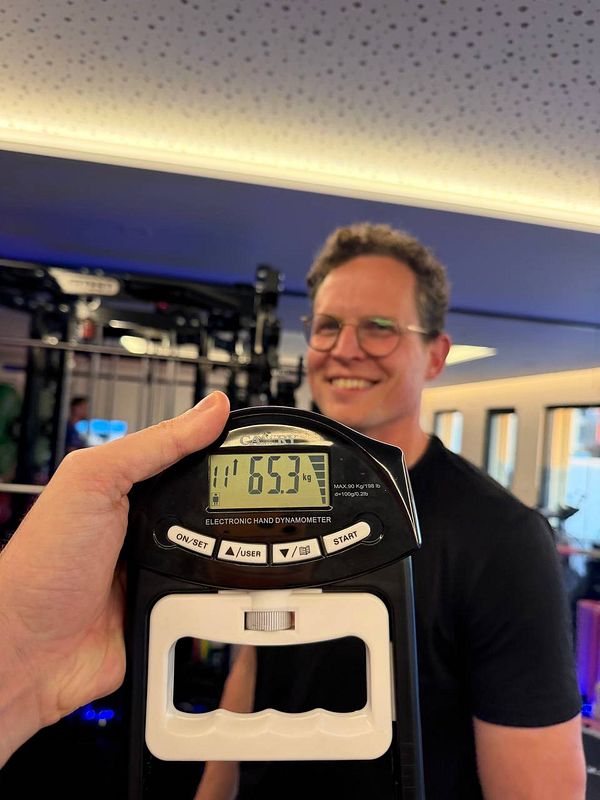 A woman in athletic wear is standing and smiling in a gym environment with workout equipment and a large mirror in the background.