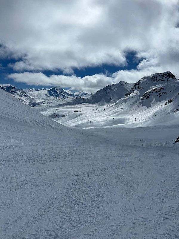 A scenic view of a snow-covered ski slope surrounded by mountains under a partly cloudy sky.