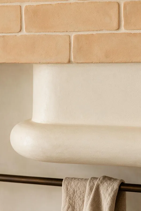 A close-up of an Organic Modern interior detail featuring a smooth cream plaster mantel with rounded edges beneath warm beige brickwork. A dark bronze rod holds a soft natural linen textile below, creating a quiet, textural contrast.
