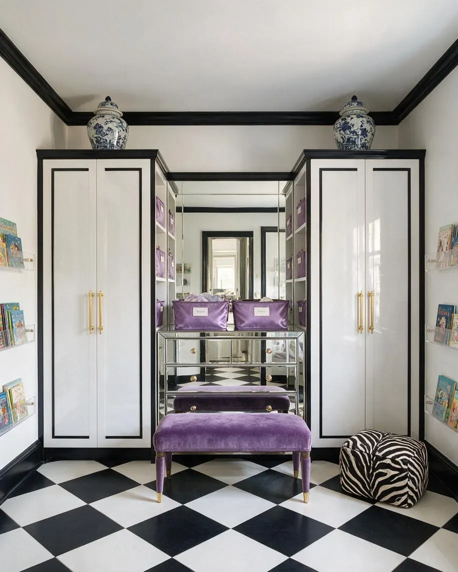 A nursery with Hollywood Regency styling features glossy white built-in cabinets outlined in black trim, a mirrored center vanity, and a black-and-white checkerboard floor. Purple velvet storage pieces and a matching bench add color, while blue-and-white porcelain jars sit on top of the cabinets and clear wall shelves display children’s books.