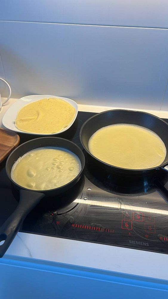 Three frying pans on a stovetop with crepe batter cooking and a plate of finished crepes.