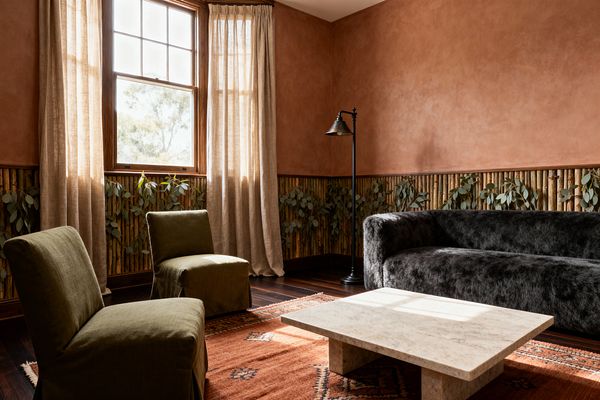 Sunlit living room with terracotta plaster walls above a bamboo wainscot threaded with hanging eucalyptus. A low rectangular travertine coffee table rests on a rust patterned rug, facing a rounded charcoal shearling-style sofa and two olive linen slipper chairs. Sheer beige curtains frame a wood-trimmed window, with a black metal floor lamp beside the sofa. Dark hardwood floors and soft daylight create a warm, organic atmosphere blending modern and rustic elements.