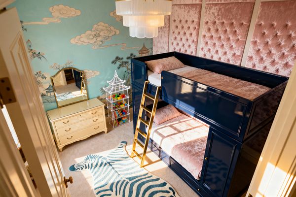 A children’s room with glossy navy built-in bunk beds has a gold ladder and blush-pink crushed-velvet bedding set before tall tufted velvet wall panels. A robin’s-egg blue Chinoiserie mural with clouds and pagodas covers the opposite wall beside a cream dresser with a gold-framed mirror and a clear pagoda-shaped shelf holding toys. A tiered frosted-glass chandelier hangs above a beige carpet with a teal-and-white zebra rug.