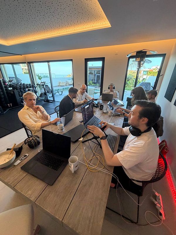 A group of seven people working together around a large table with laptops in a modern indoor coworking space with large windows overlooking an outdoor area.
