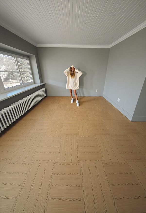 A woman stands in a spacious, empty room with a light gray wall and wooden flooring.