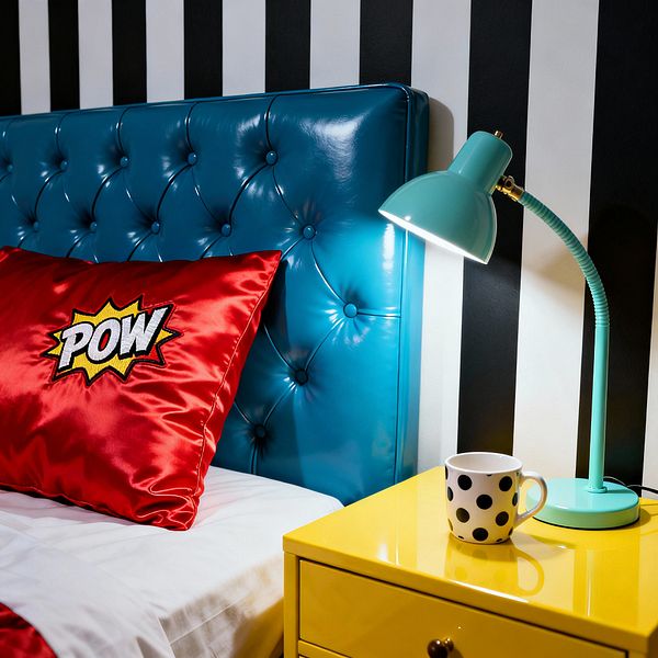 A bedroom vignette in Pop Art style shows a glossy teal, button-tufted headboard against bold black-and-white vertical striped wallpaper. A red satin pillow embroidered with a comic 'POW' graphic sits on white bedding beside a bright yellow lacquered nightstand topped with a mint metal task lamp and a white mug with black polka dots, all illuminated by the lamp's focused light.
