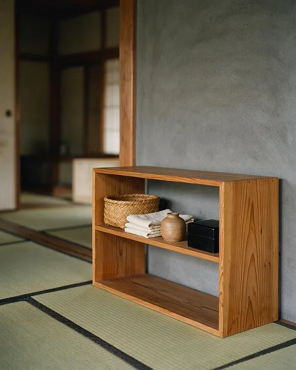 A simple low wooden shelving unit sits beside a textured gray wall in a Japanese-style room with tatami mats. The shelf holds a woven basket, folded white linens, a small ceramic vase, and a black box, creating a calm, uncluttered Zen composition.
